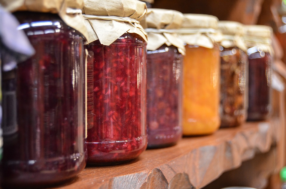 6 jam jars lined up on a shelf. Jars filled with preserves and sealed with wax paper and string