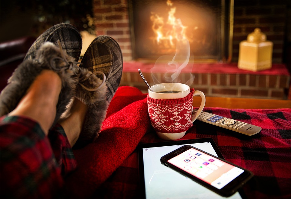 person with feet up with mobile phone and tablet next to them as well as a steaming cup of hot drink before a fire